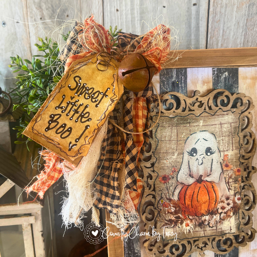 Close up of Whimsical Ghost with Pumpkin Wood Canvas Decor showing hand-lettered Sweet Little Boo distressed tag layered black buffalo check orange crinkle ribbon wispy raffia bow and rusty jingle bell by Country Charm by Tracy