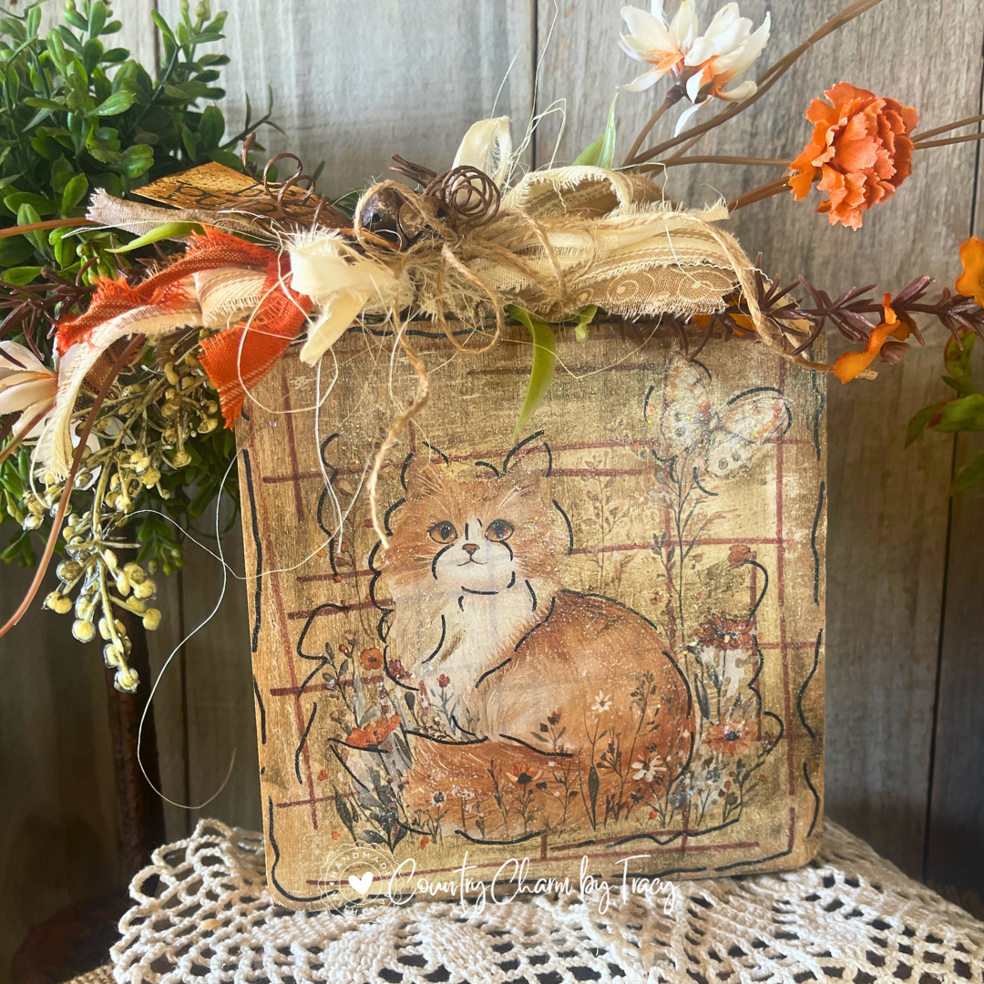 Close up of Hey Pumpkin Fluffy Orange Cat Wood Block Decor showing hand-lettered hey pumpkin distressed tag rusty jingle bell cluster layered cream lace and orange ribbon bow with wispy raffia by Country Charm by Tracy
