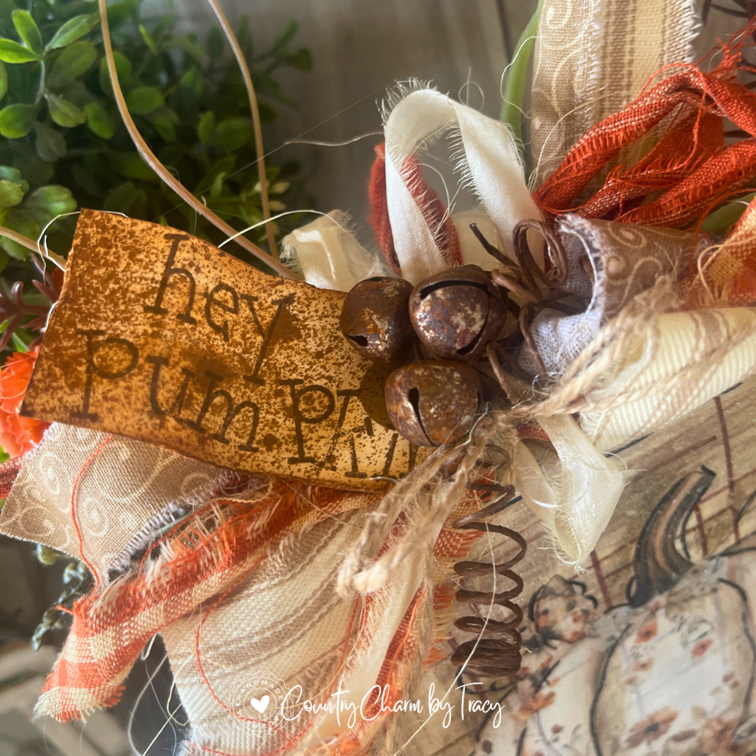 Close up of Hey Pumpkin Floral Pumpkin Wood Block Decor showing hand-lettered hey pumpkin distressed tag rusty jingle bell cluster curly wire spring and layered orange buffalo check cream ribbon bow with wispy raffia by Country Charm by Tracy